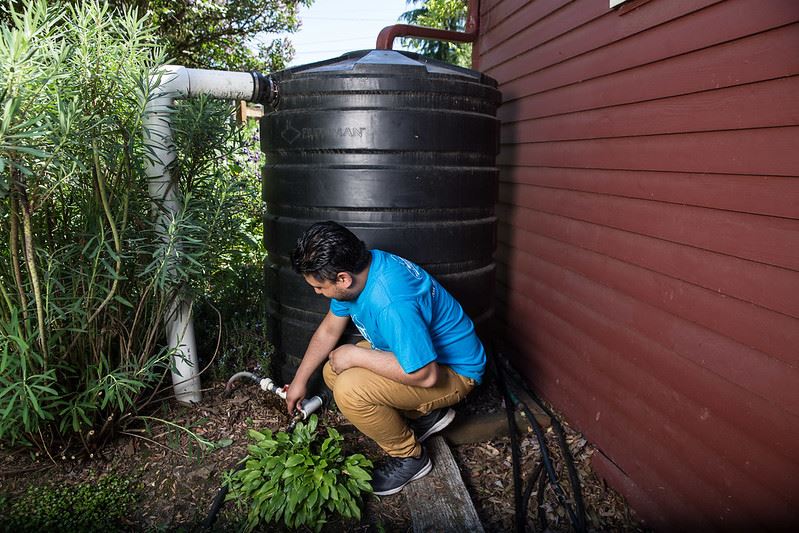 Rain barrel on side of home with person in foreground bending down to touch a valve.